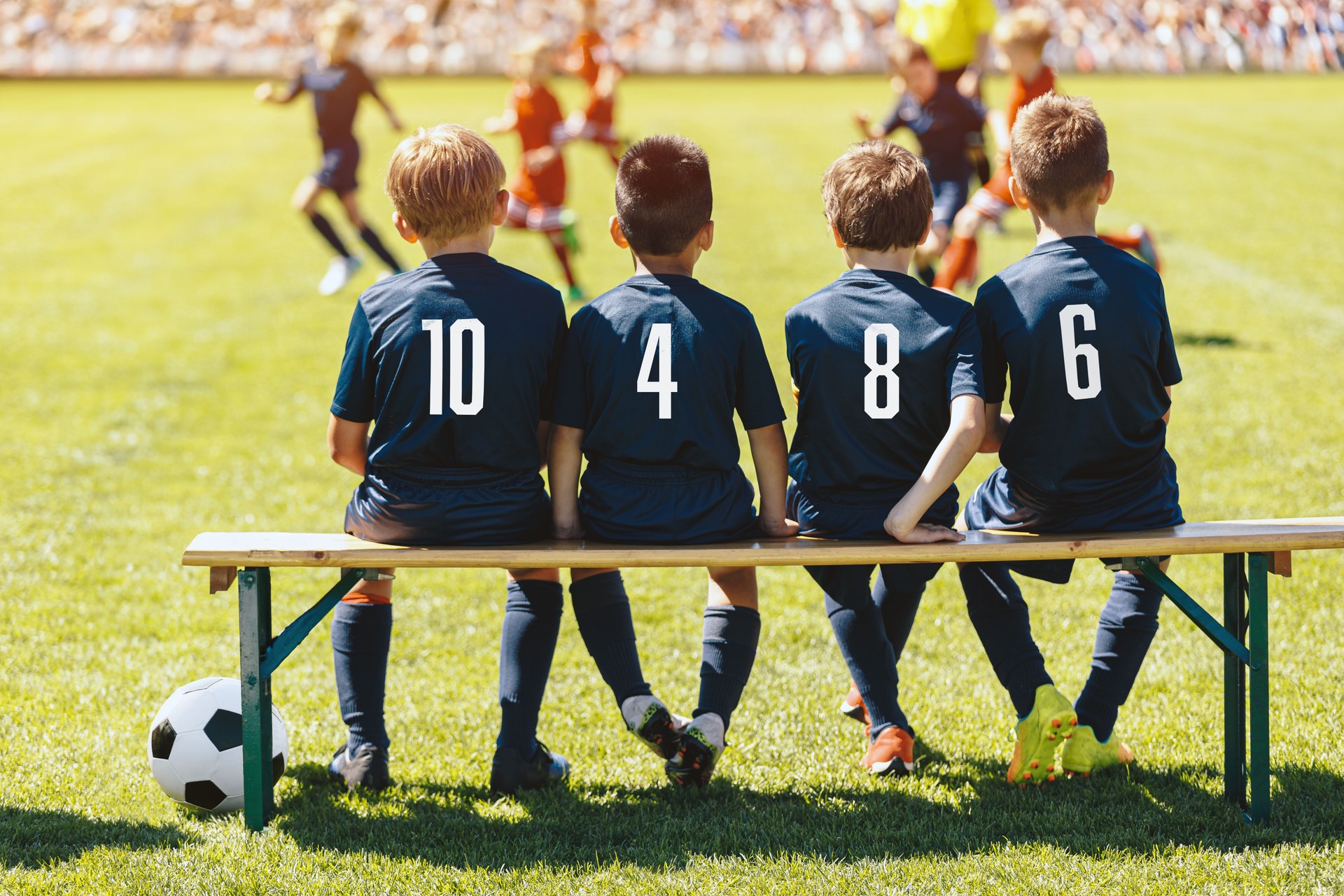Ersatzspieler in der Fußballmannschaft sitzen auf der Bank. Multiethnische Gruppe von Kindern in einer Sportmannschaft unterstützt eine Fußballmannschaft in einem Spiel. Mitglieder der Schulfußballmannschaft in blauen Trikots mit Nummern